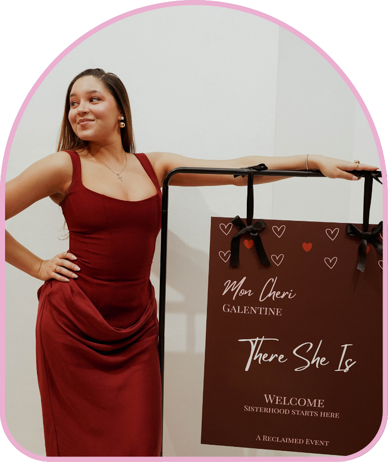 Woman in a red gown standing next to a sign with the text 'Mon Cheri GALENTINE There She Is WELCOME SISTERHOOD STARTS HERE A RECLAIMED EVENT' decorated with small heart graphics.