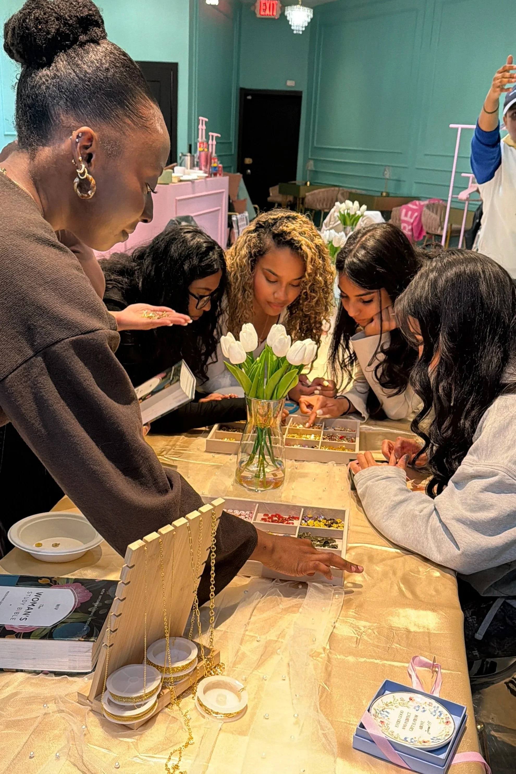A group of women gathered around a table, closely examining jewelry and accessories. The table has a vase of white tulips and various jewelry items for sale, with decorative display stands and boxes. The setting appears to be an indoor shop or boutique with pastel-colored walls and elegant decor.