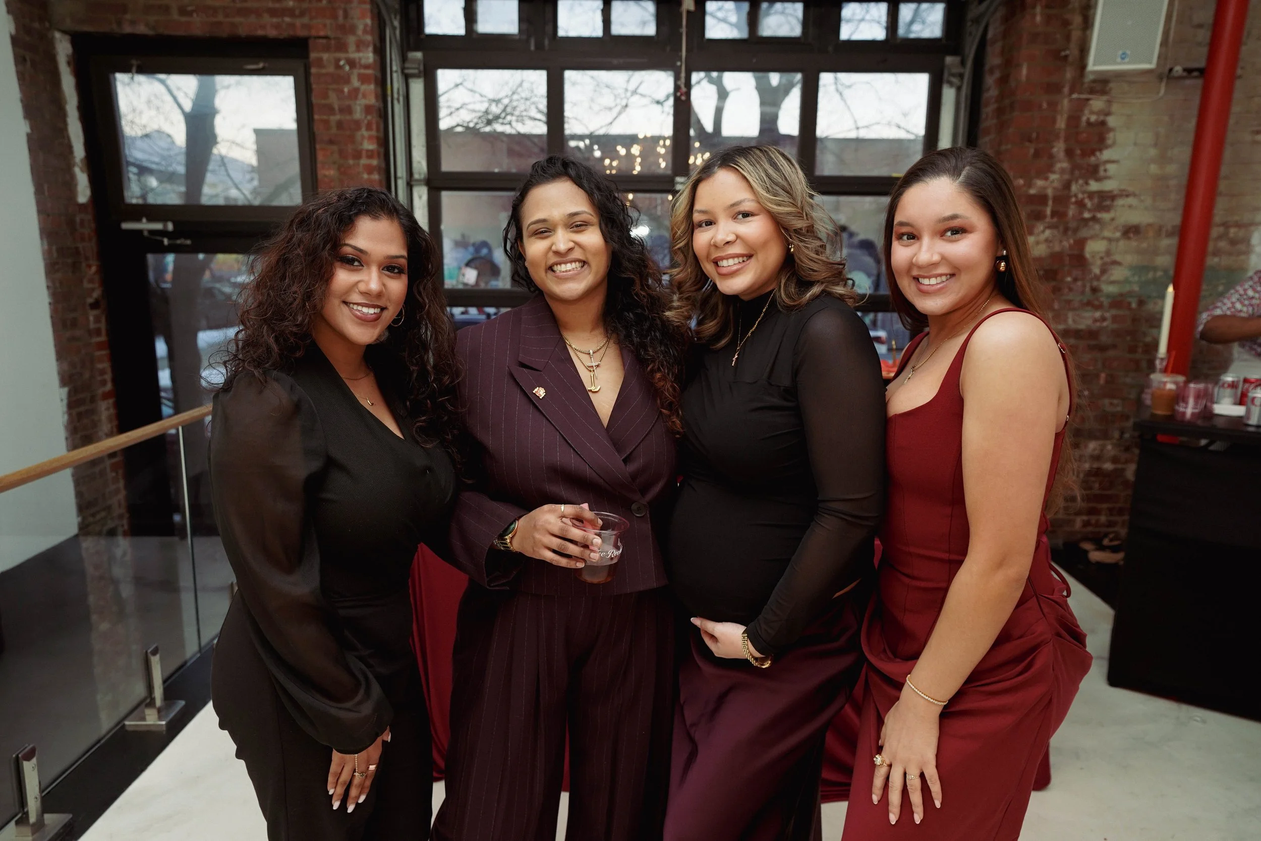Four women standing together indoors, smiling for a photo. They are dressed in formal or semi-formal attire, with three wearing dark colors and one in a maroon dress. Behind them, there are large windows with a view of trees and outdoor lights, and a brick wall.