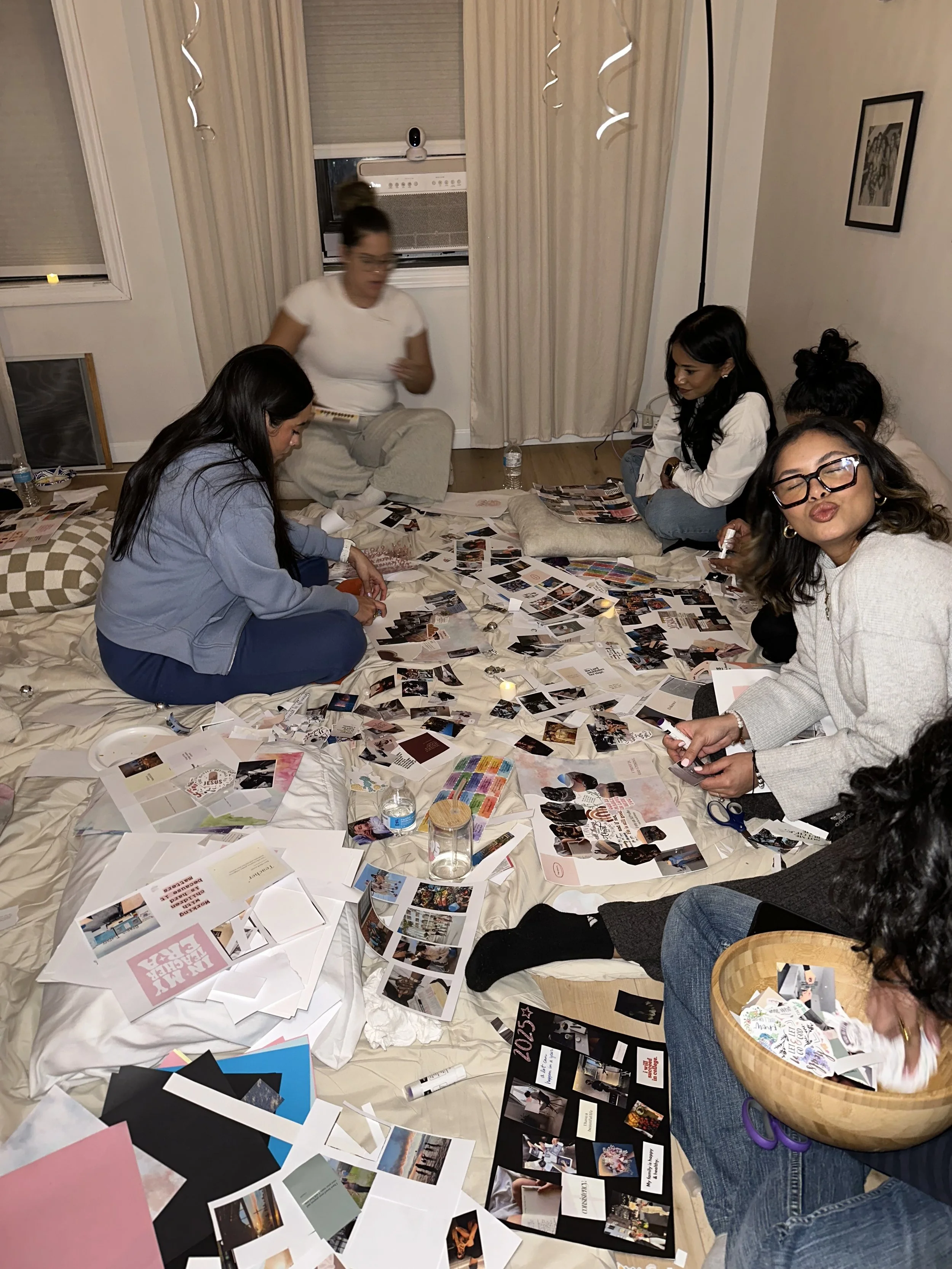 A group of women sitting on a bed in a room, surrounded by scattered photos, cards, and papers, engaging in a creative or planning activity.