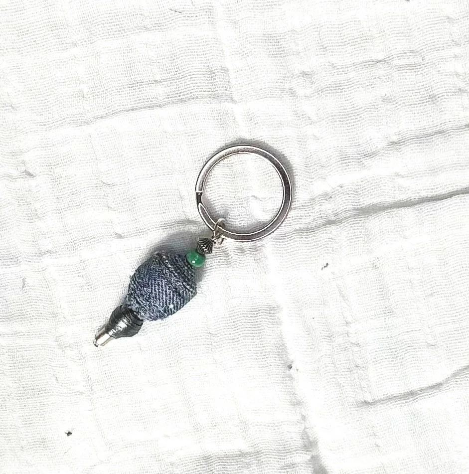 A keychain with a tiny fabric sock and a small green bead attached, lying on a white textured cloth.