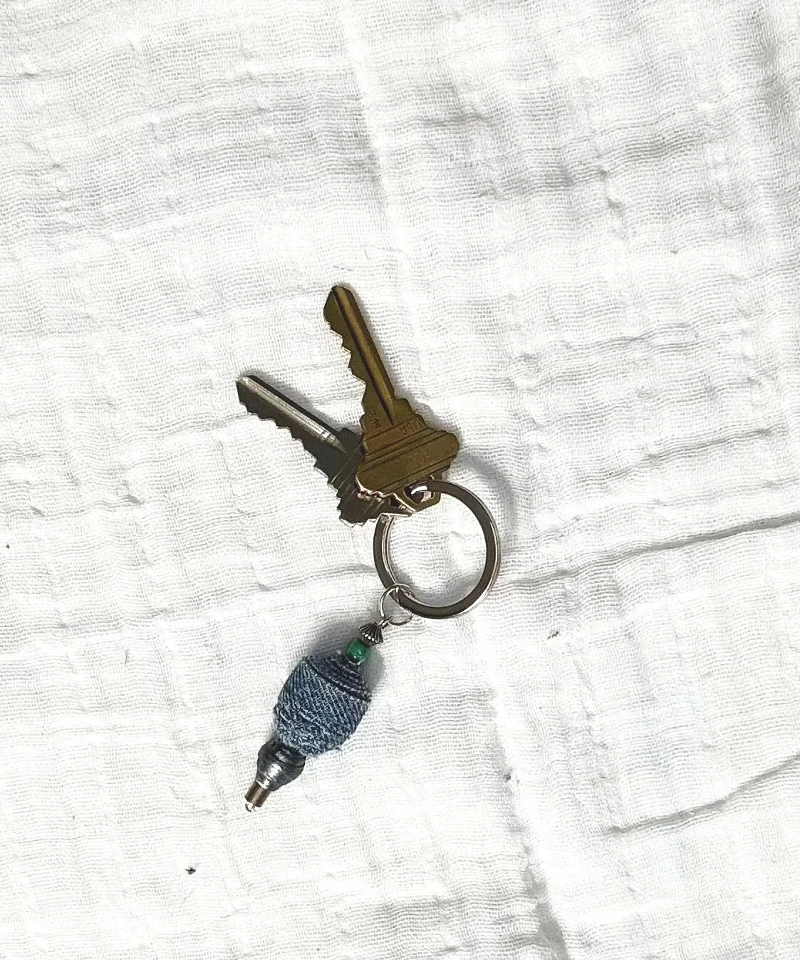 A set of two keys attached to a keyring with a decorative cloth bead and bead cap, resting on a white fabric surface.