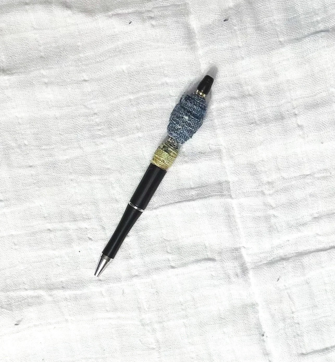 A black pen with a decorative wrap around the top part of the barrel, lying on a white textured fabric surface.