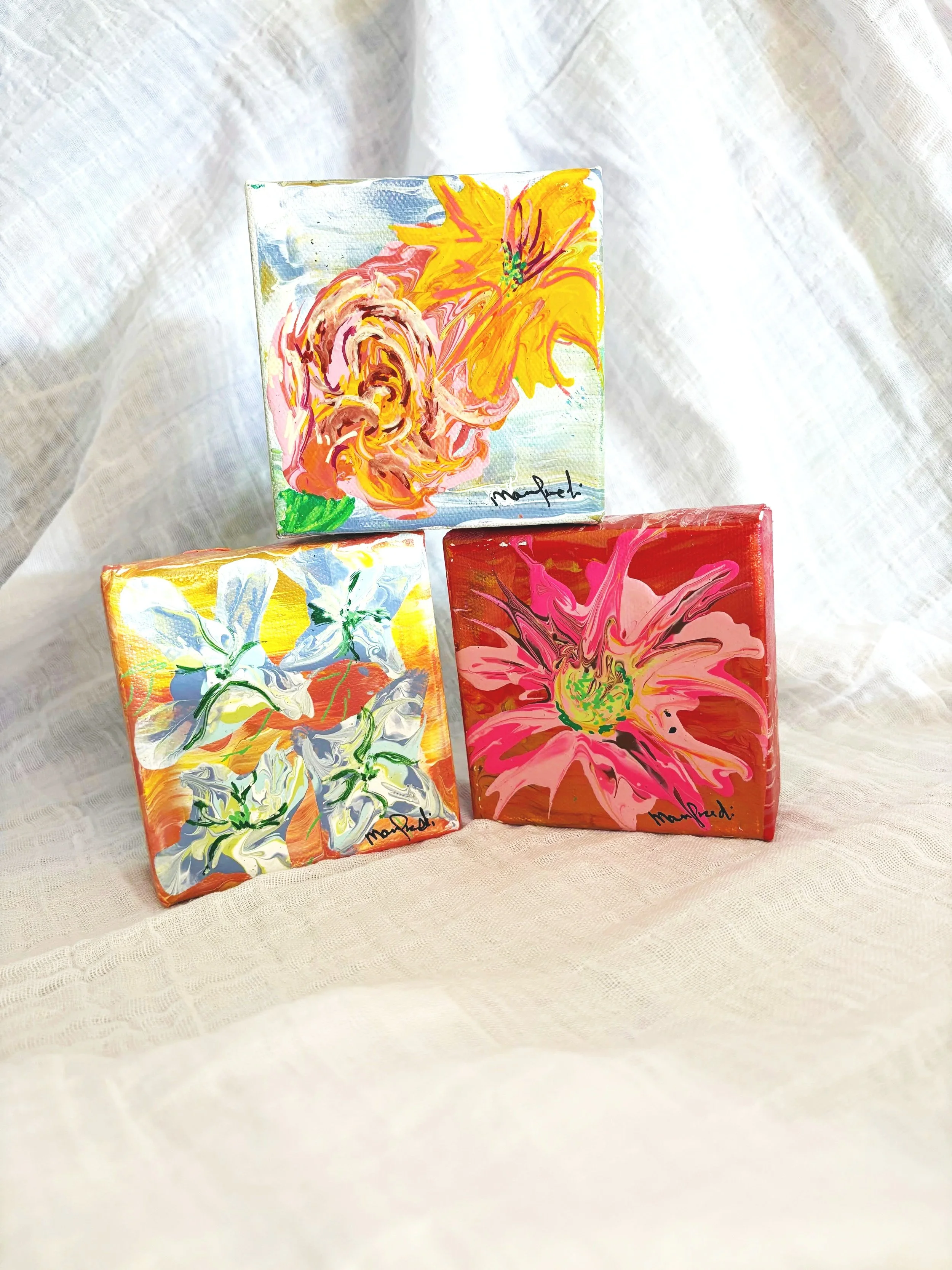 Three small canvases with colorful abstract flower paintings placed on a white textured fabric background.