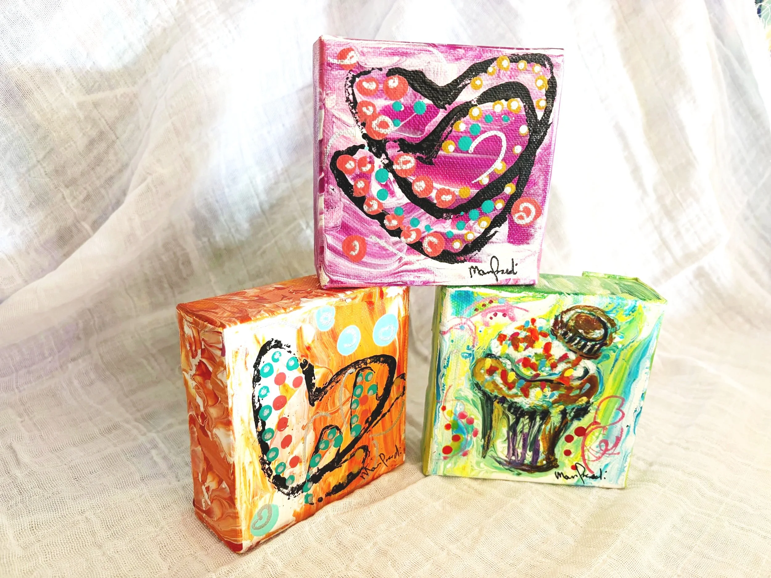 Colorful abstract paintings on small canvas blocks, featuring swirls, dots, and bold lines, displayed on a fabric surface.