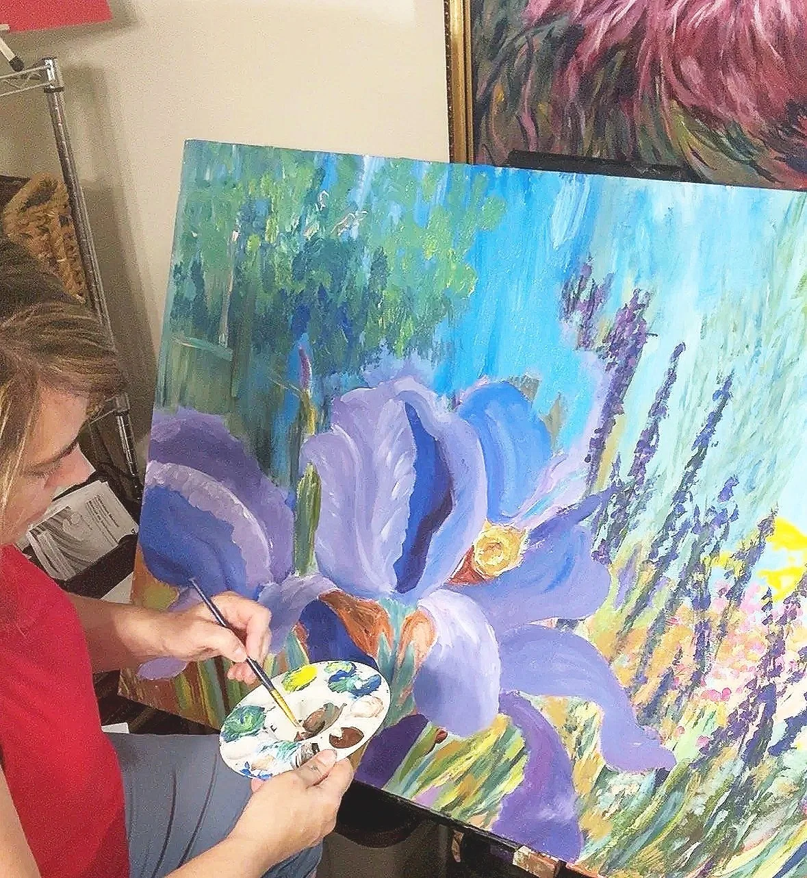 A woman painting a large, colorful floral scene on a canvas with a palette in her hand, focusing on purple and blue flowers and green foliage.