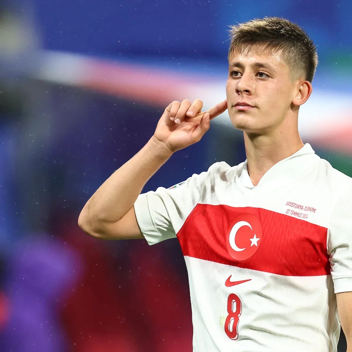 A young male soccer player wearing a Turkish national team jersey with the Turkish flag emblem, touching his ear with his right hand, looking focused.