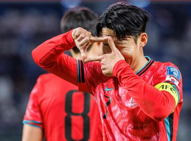 A soccer player wearing a red jersey with the number 7 making a heart shape with his hands in front of his face, celebrating on the field with another player in the background.