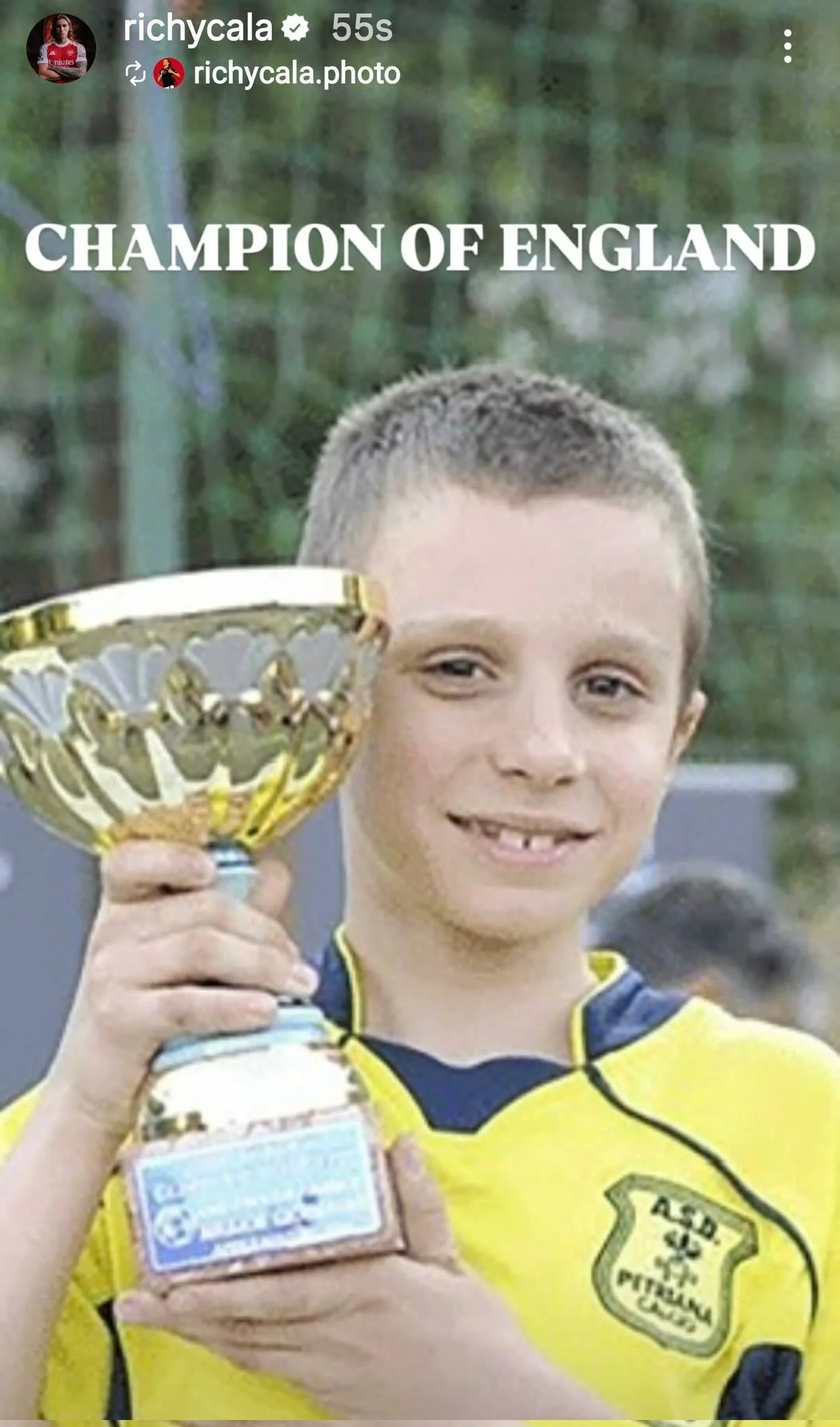 A young boy in a yellow and blue sports jersey holding a trophy and a medal, smiling. The jersey has a badge that reads 'ASD PUGLIA.' The background is blurry with trees.