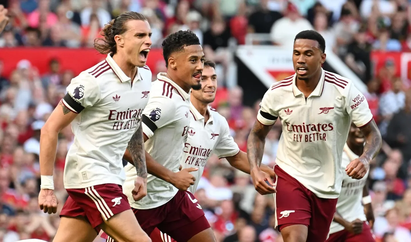 Four soccer players celebrating on the field, wearing white jerseys with maroon shorts, with a crowd of spectators in the background.