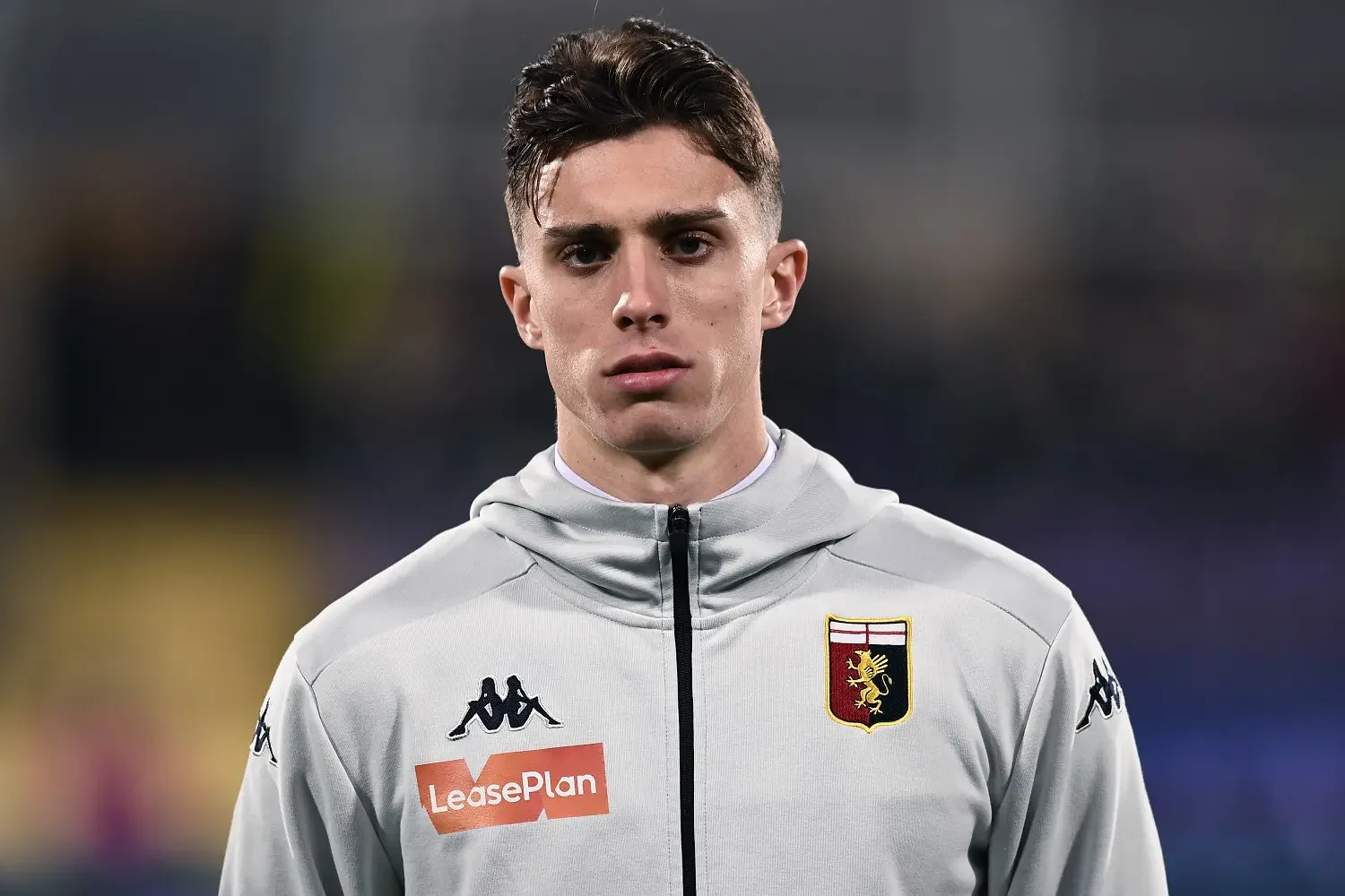 A young male soccer player with short dark hair, wearing a light gray jacket with the Genoa football club crest and sponsorship logos, standing outdoors with a blurred background.