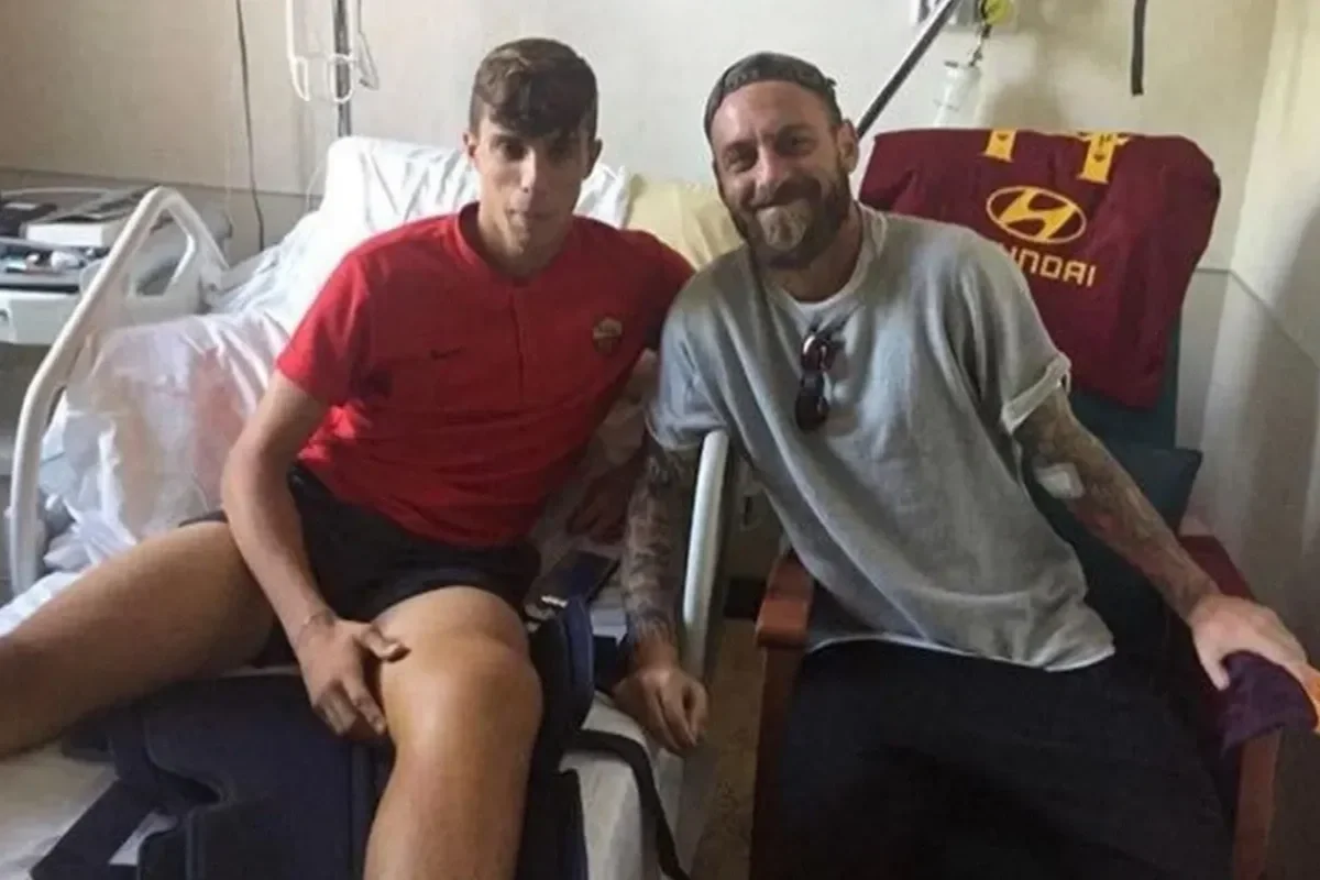 A young man and a middle-aged man sitting together in a hospital room, smiling at the camera. The young man is wearing a red shirt and black shorts, sitting on the edge of a hospital bed. The middle-aged man is wearing a gray t-shirt, with tattoos on his arms, and sunglasses hanging from his shirt collar, sitting on a chair beside him.