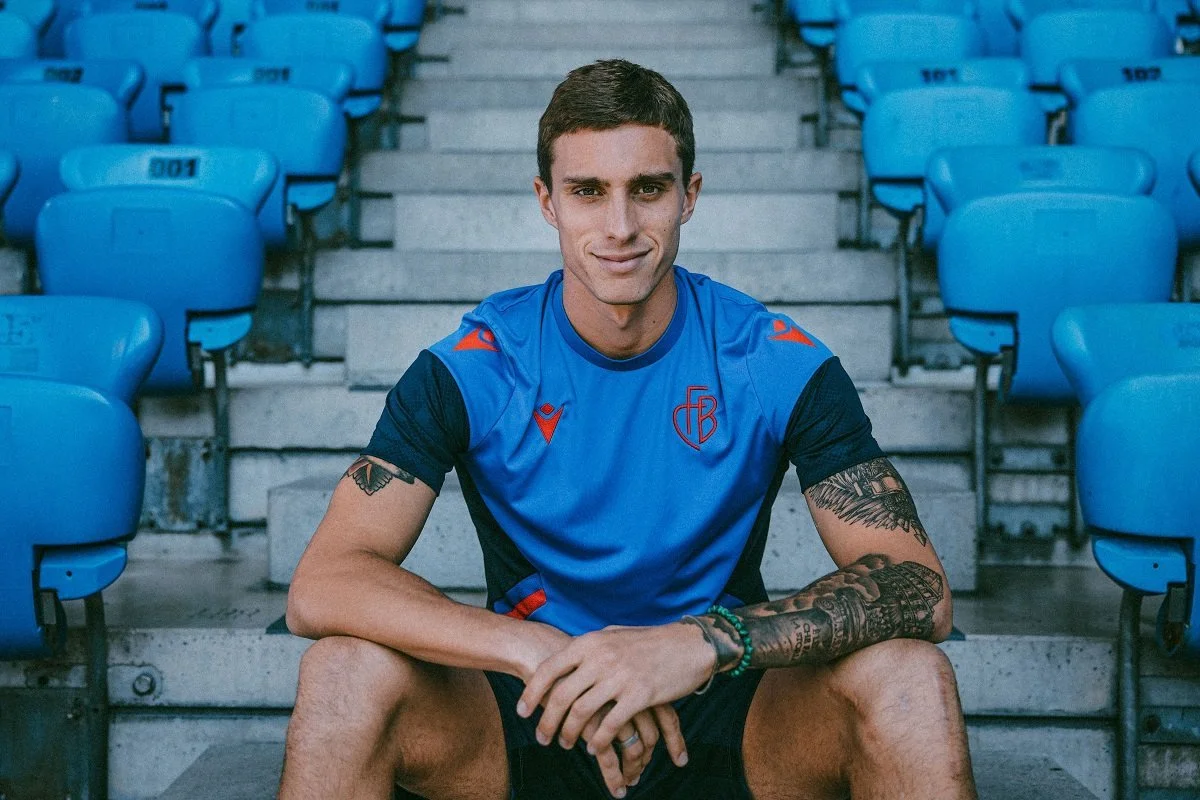 A young man with tattoos on his arms, wearing a blue sports jersey, sitting on stadium bleachers with blue chairs behind him, looking at the camera.