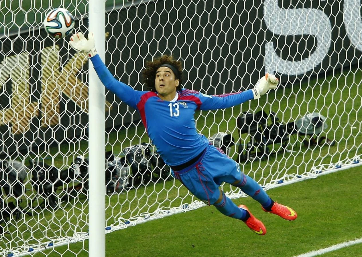 A soccer goalkeeper in a blue uniform with the number 13 is diving to catch a ball in the goal during a game.