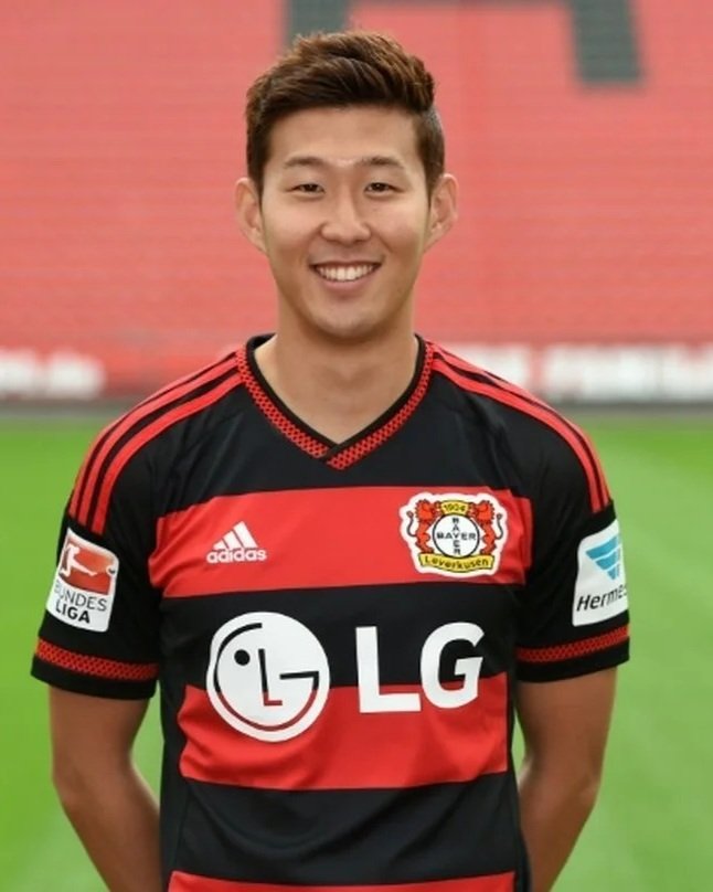 Young man smiling, wearing a black and red soccer jersey with the Bayern Leverkusen logo, on a soccer field.