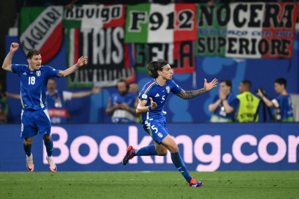 Italian female soccer player celebrating a goal on the field, wearing a blue uniform with the number 5, with a male teammate in the background also celebrating, and banners with Italian text and colors in the background.