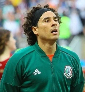 A male soccer player wearing a green jacket with the Mexico national team emblem, standing on a field with a focused expression.