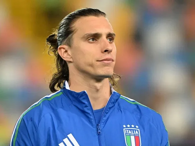 A man with long hair tied back, wearing a blue sports jacket with the Italian national team logo and stars, standing outdoors with a blurred audience in the background.