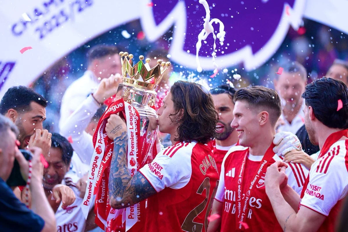 Soccer players celebrating with a trophy, one player kissing the trophy, surrounded by confetti and teammates in an outdoor setting.