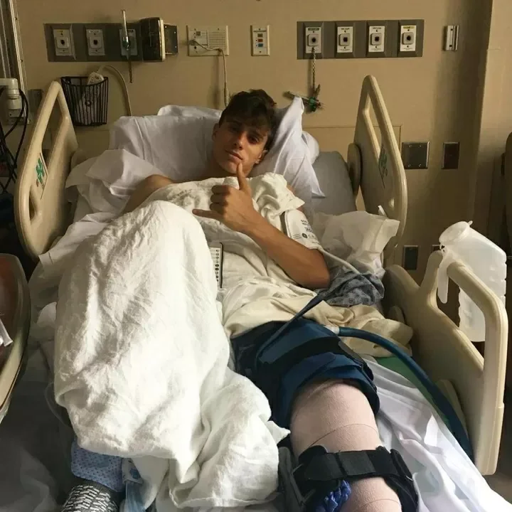 Young man lying in a hospital bed with medical equipment and monitors in the background, giving a thumbs-up.