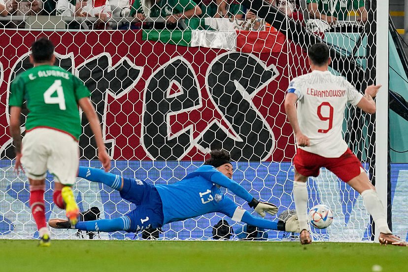 A soccer player in a white and red uniform kicks a ball toward the goal as two opposing players attempt to block it. The goalkeeper in blue dives to the side in an effort to save the shot. The scene is on a soccer field with a net in the background and spectators watching from the stands.