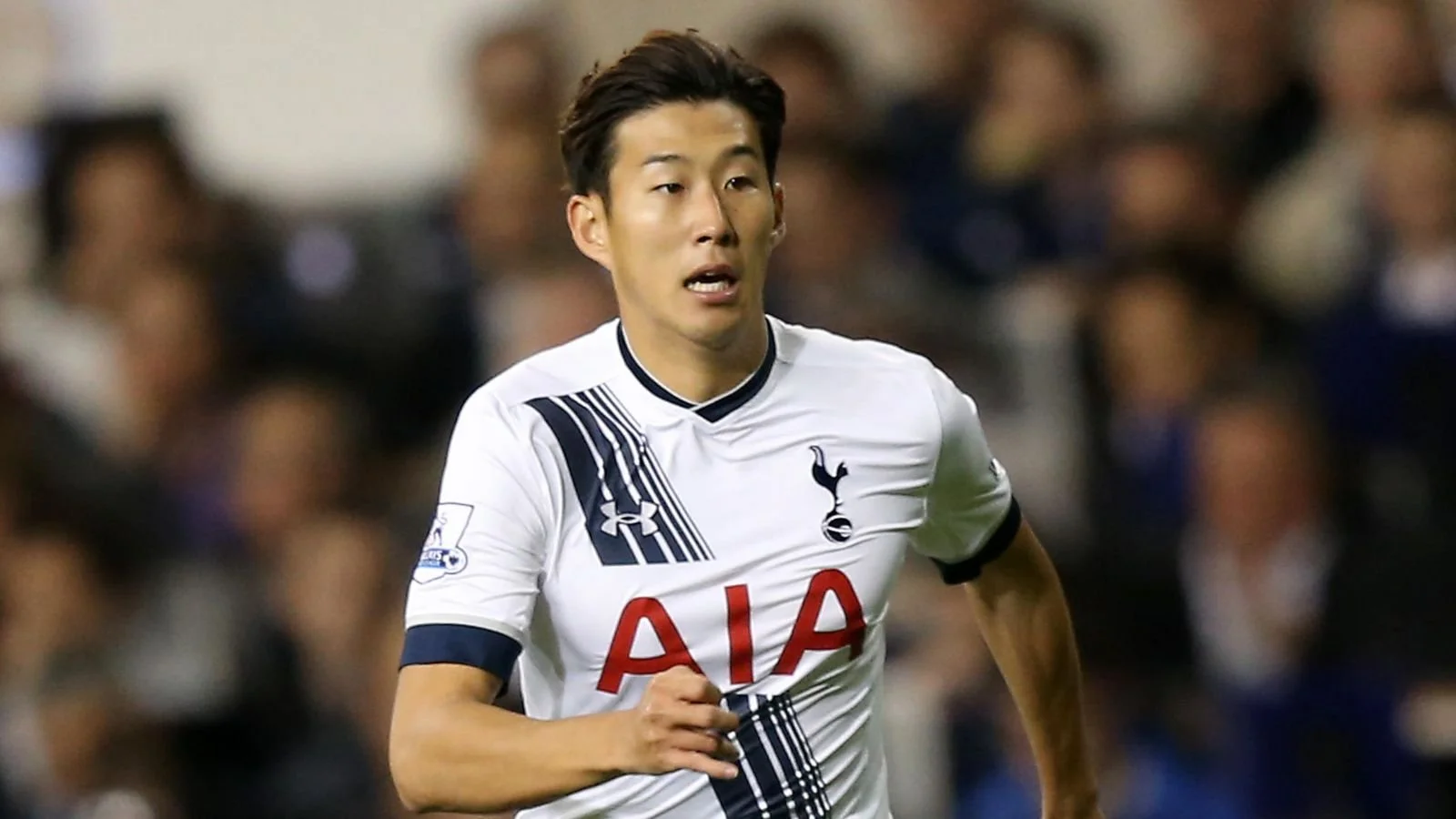 A soccer player in a white Tottenham Hotspur jersey running on the field during a match, with blurred spectators in the background.