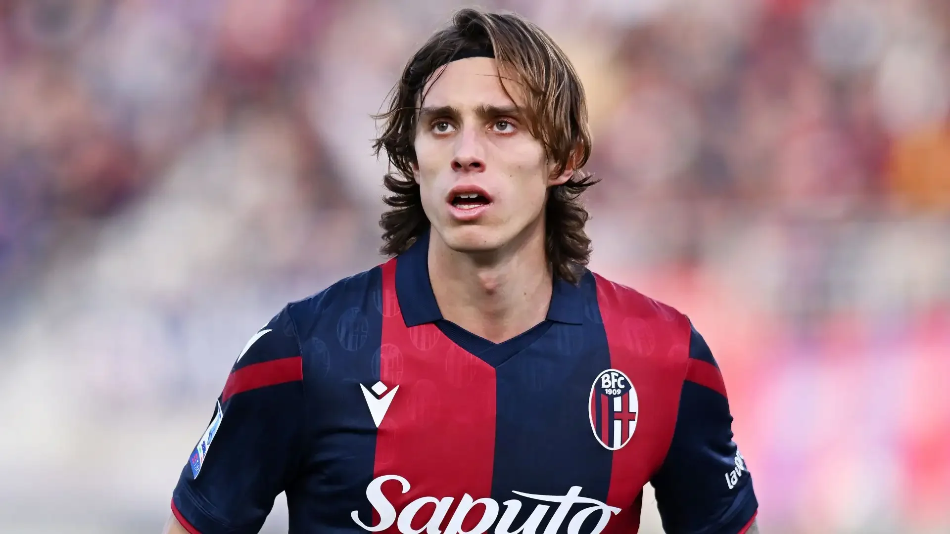 A male soccer player with long brown hair, wearing a red and blue striped jersey of Bologna FC, looking focused on the game.