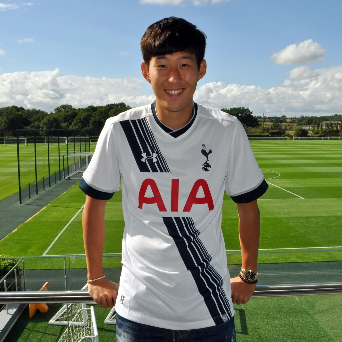 Young man smiling at the camera, wearing a white Tottenham Hotspur soccer jersey, standing outdoors on a sunny day with a soccer field and goal in the background.
