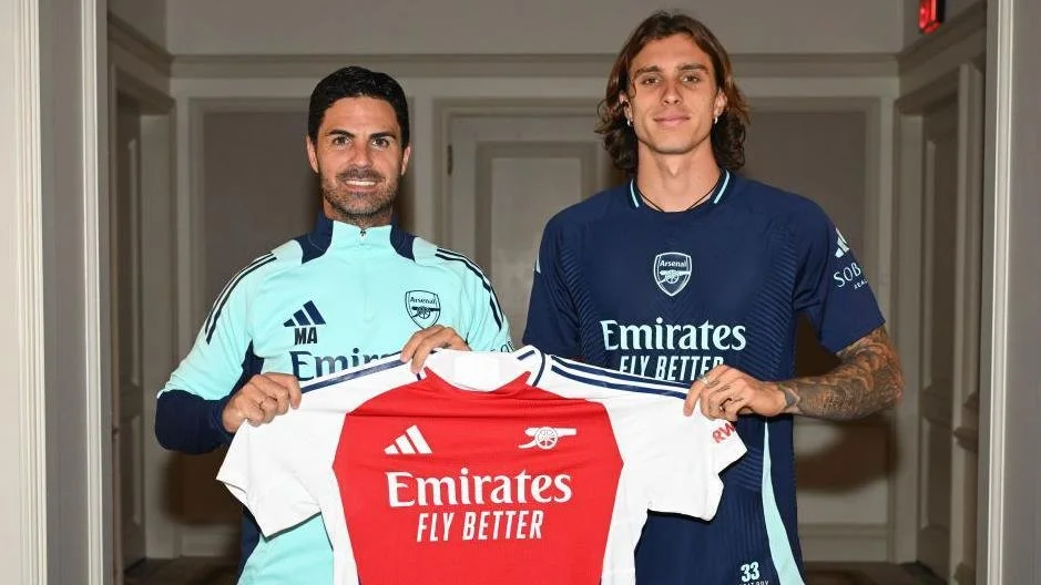 Two men holding an Arsenal football jersey together, one wearing a light blue jersey and the other wearing a dark blue jersey, standing inside a hallway.