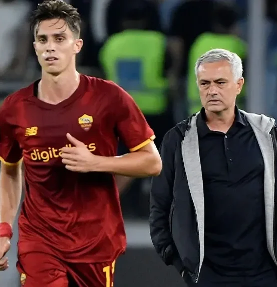 A young male soccer player in a red AS Roma jersey walking alongside an older man with gray hair, wearing a black shirt and gray jacket, on a soccer field.
