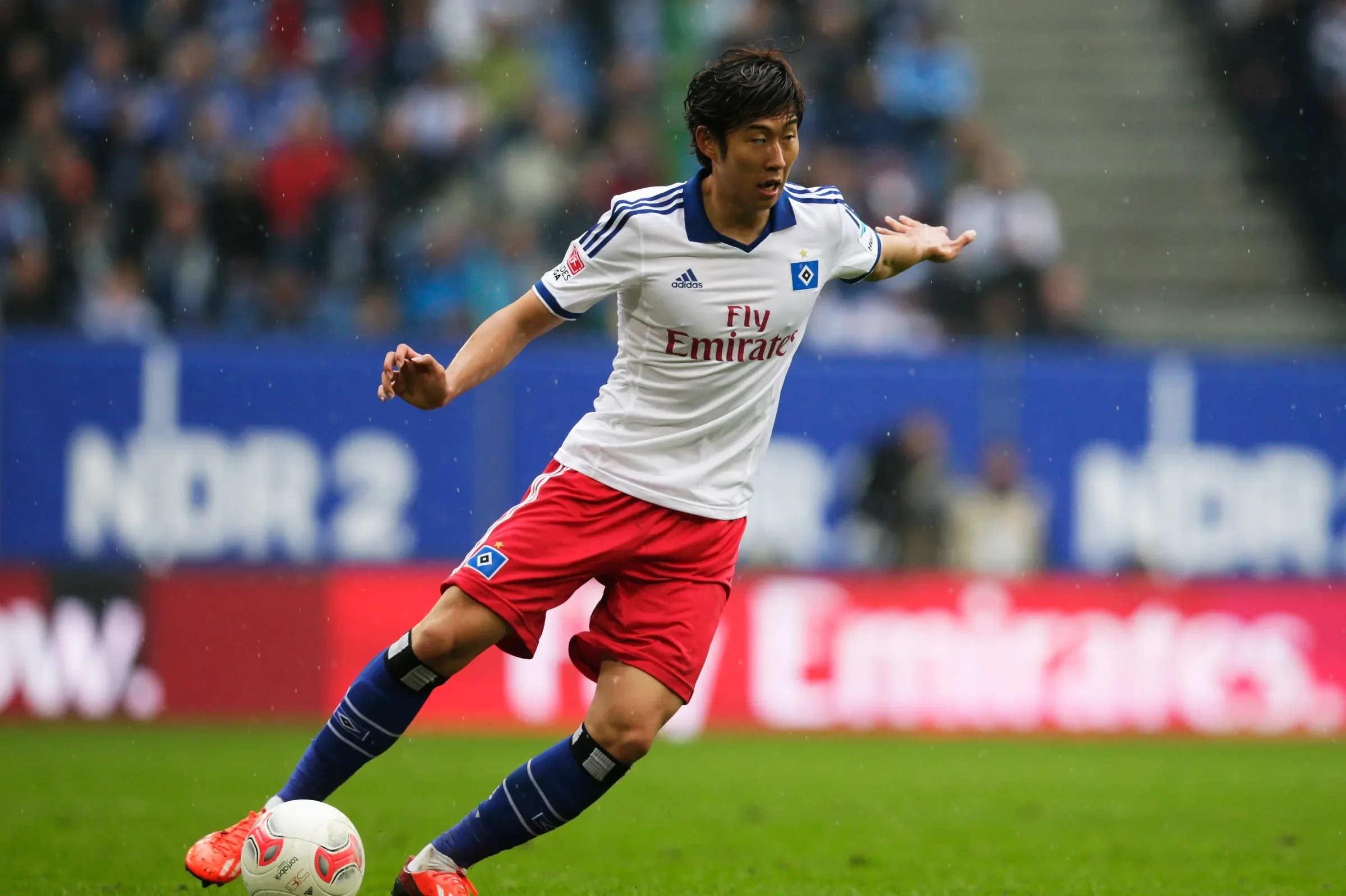 A soccer player wearing a white and blue jersey, red shorts, and red cleats, is running with a soccer ball on a grass field during a game under rainy weather.