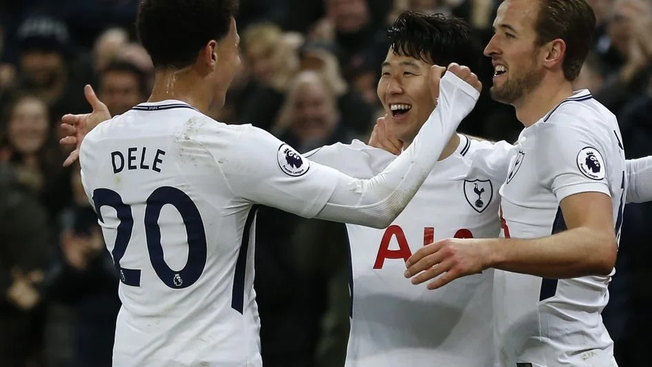 Three soccer players celebrating on the field, two in white jerseys with Tottenham Hotspur logo, one in a white jersey with Arsenal logo, in front of cheering crowd.