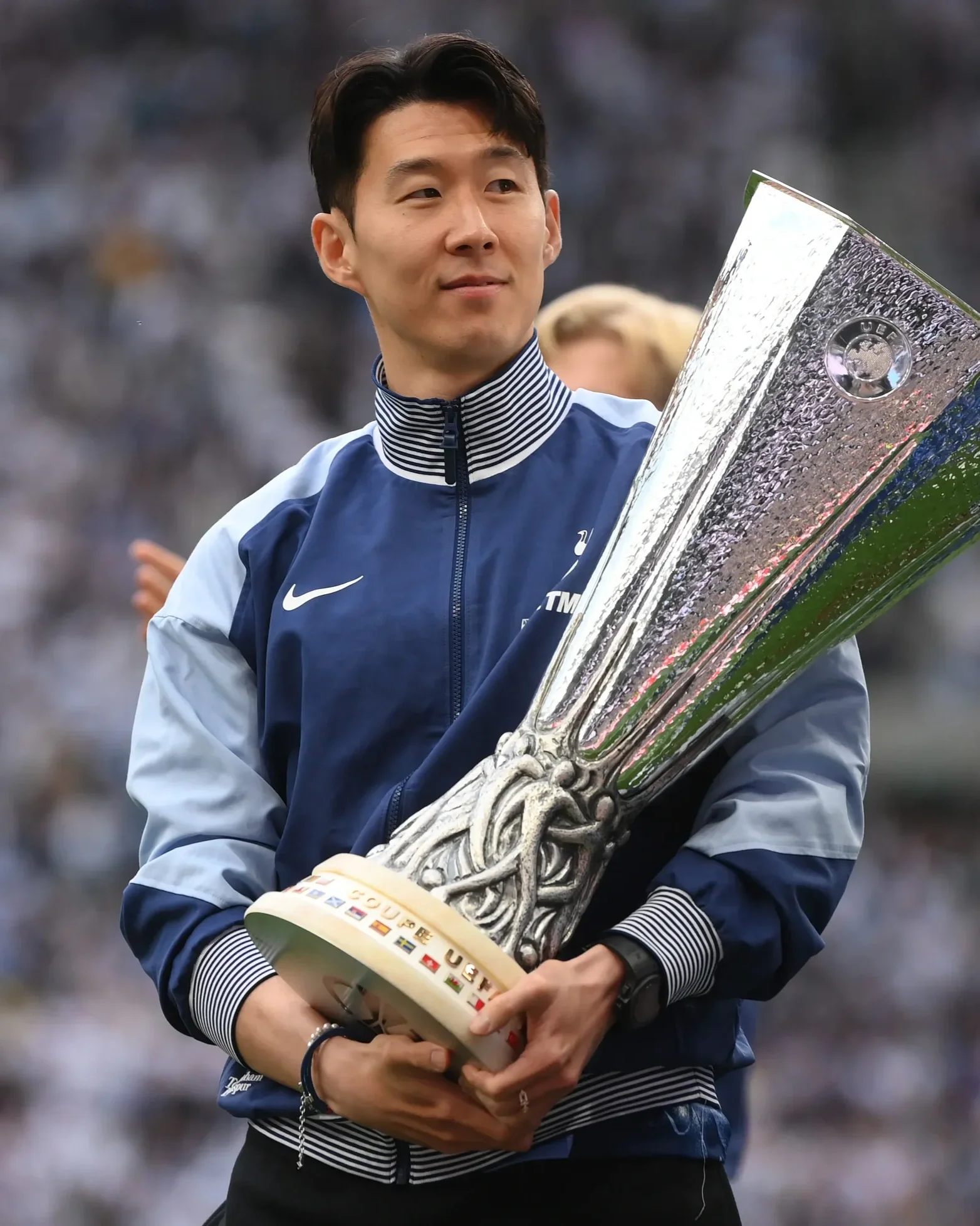 A man holding a large shiny silver trophy with a black base, wearing a blue and light gray sports jacket, in a stadium setting.