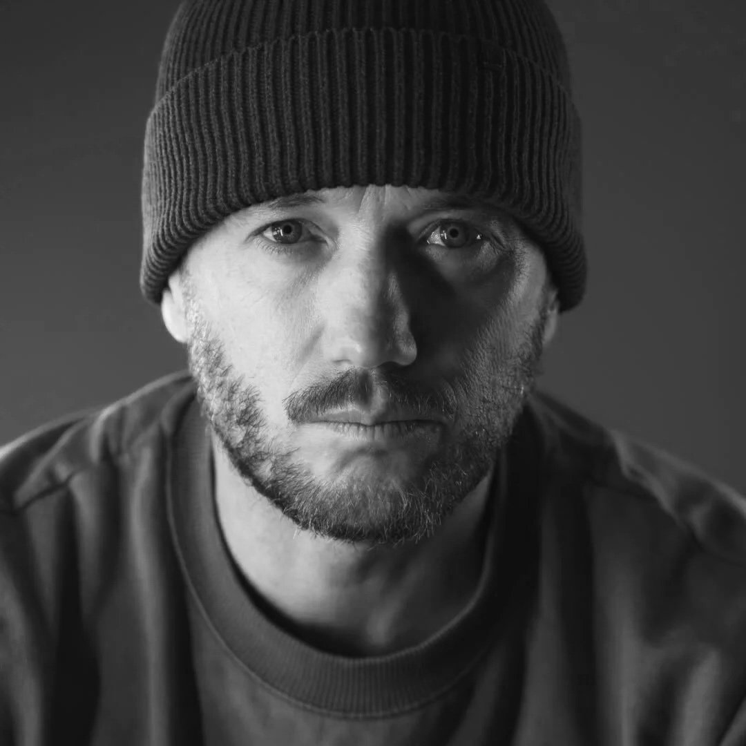Black and white portrait of a man with a beard, wearing a knit beanie, staring directly at the camera.