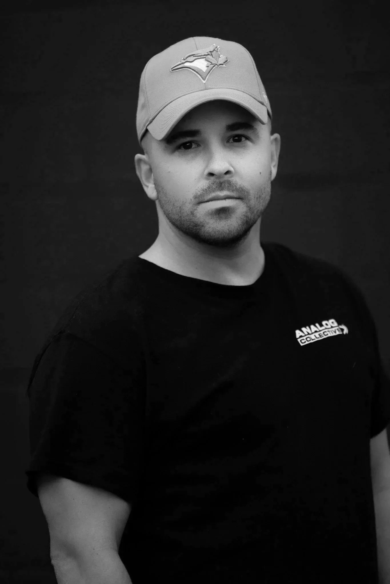 Black and white photo of a man wearing a baseball cap with a Toronto Blue Jays logo, black T-shirt with "Analog Collective" text, against a dark background.