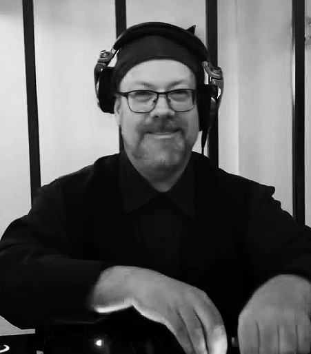 A man with glasses, a beard, and a mustache wearing headphones and a black shirt, smiling at the camera.