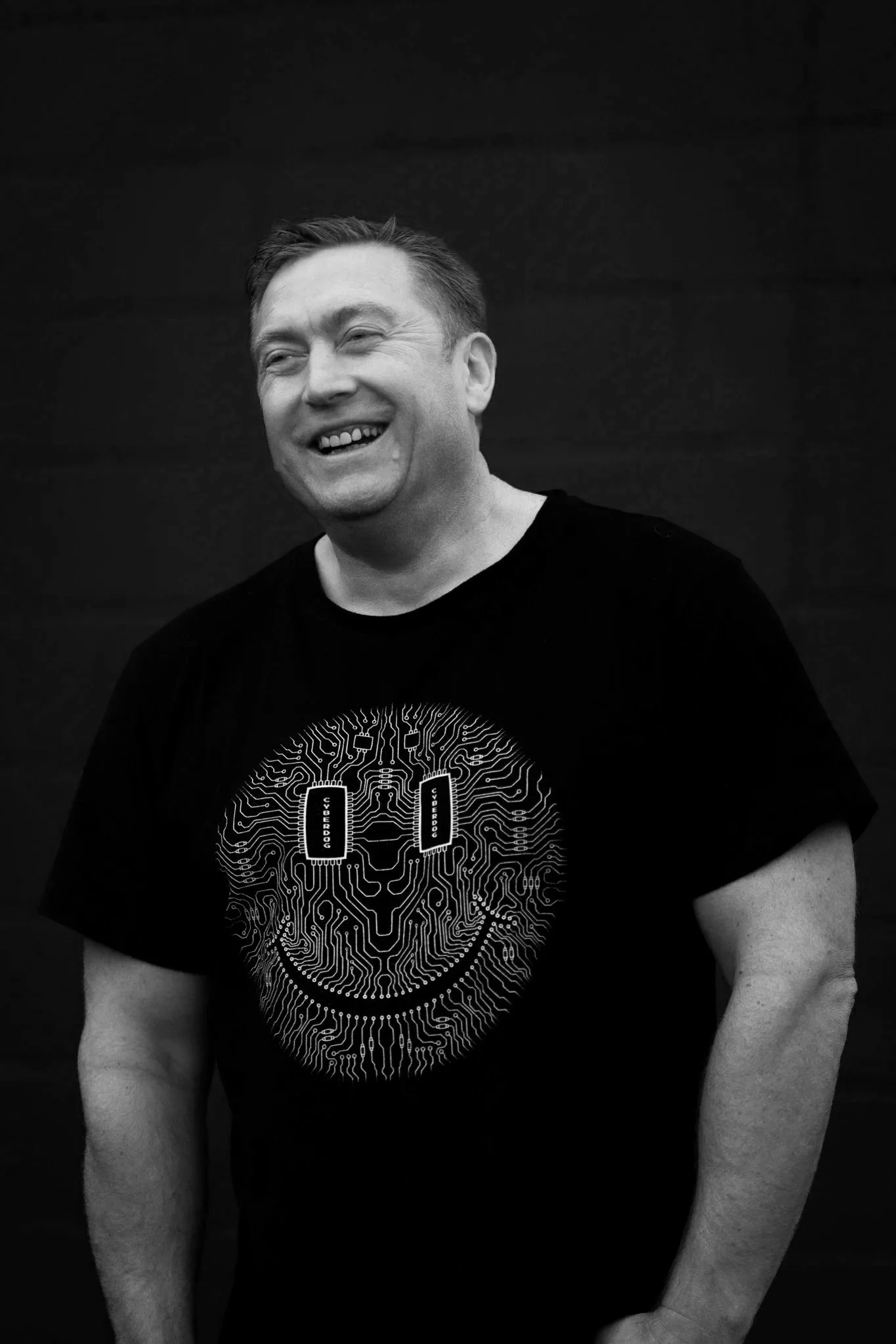 A man wearing a black T-shirt with a circuit-themed design that includes the words 'Cyber Dog'. He is smiling and looking to his left, standing against a dark background.