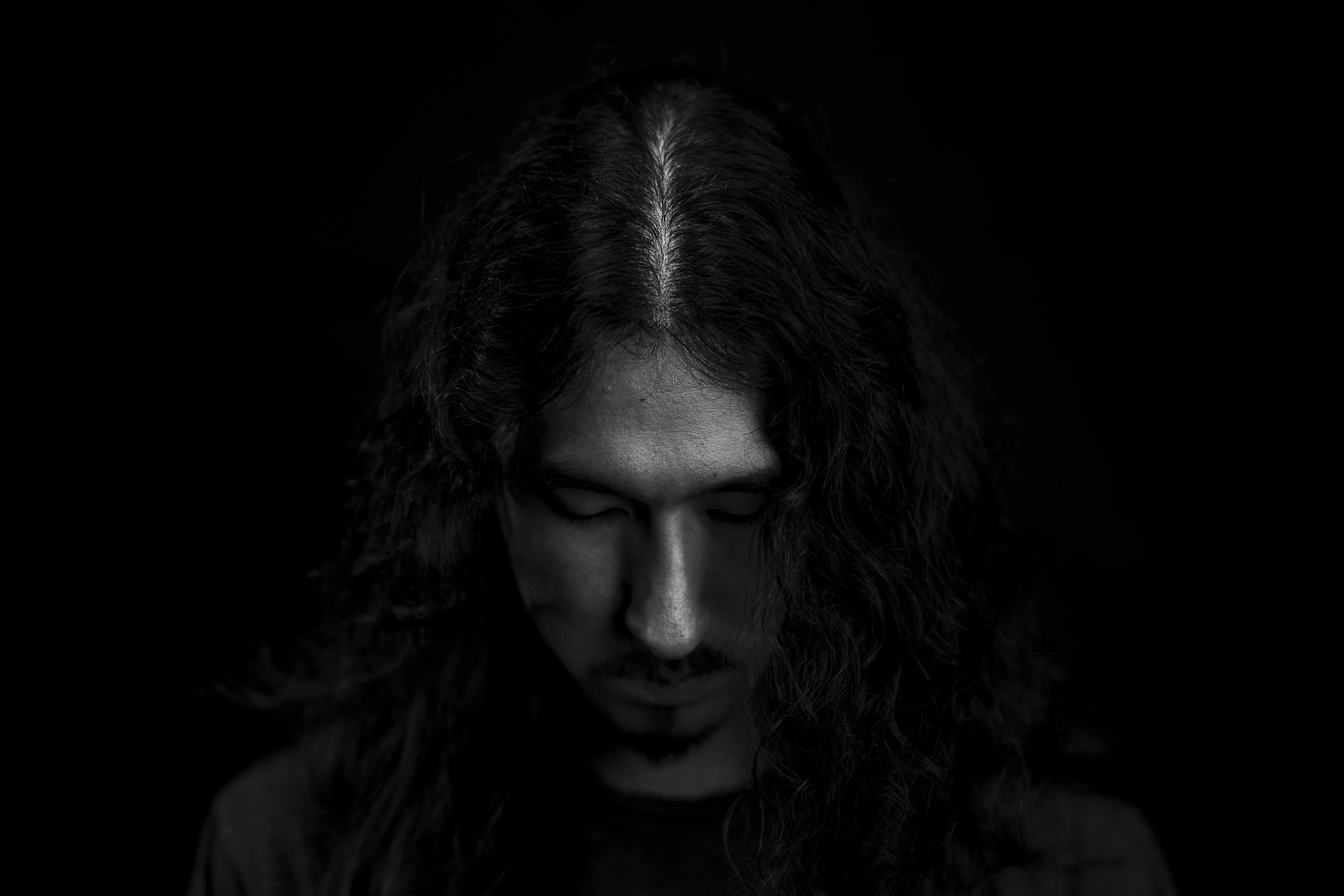 Black and white photo of a man's head, with the top of her face and hair visible, looking downward on a dark background.