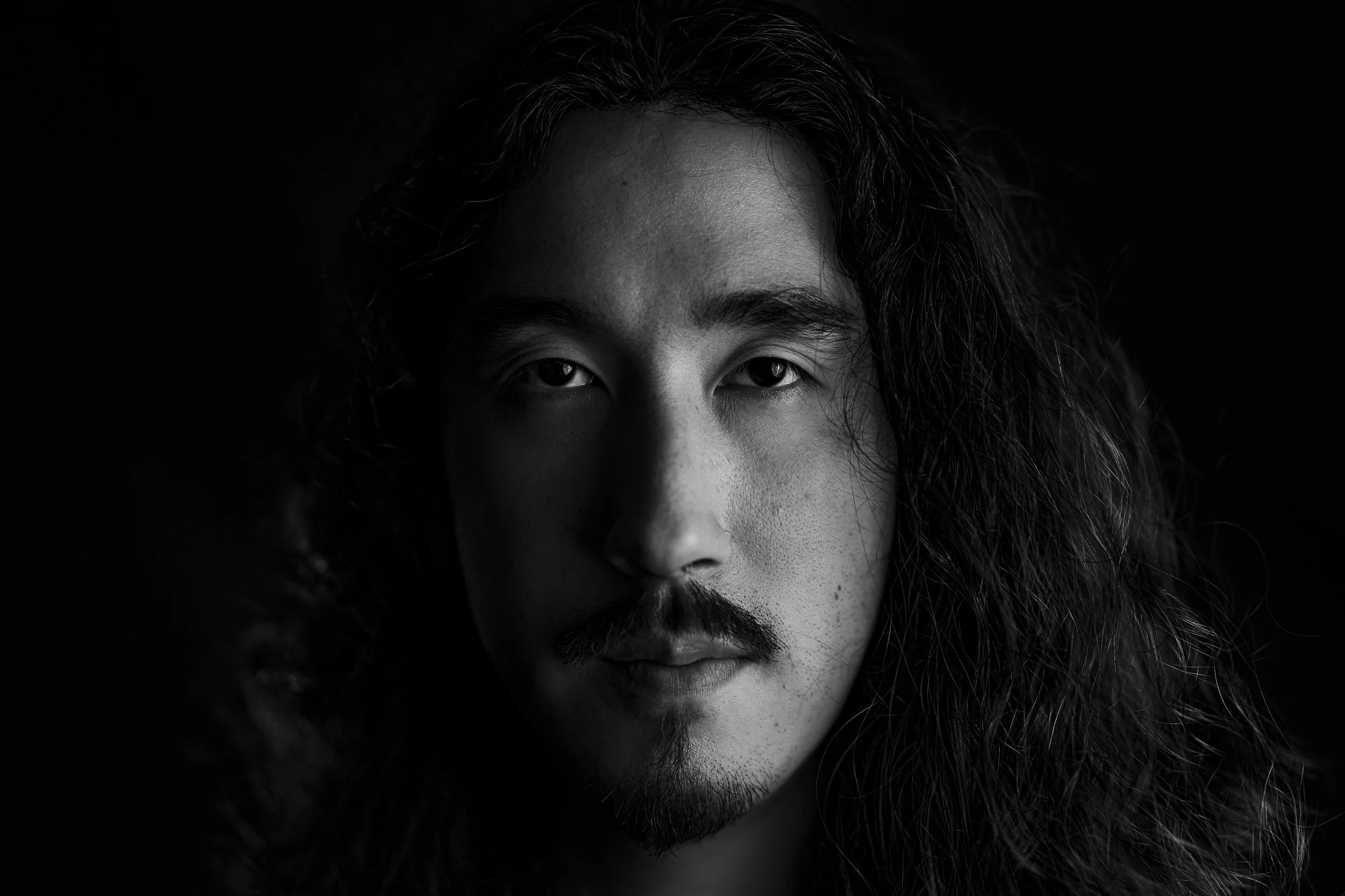 Close-up black-and-white portrait of a man with long, wavy hair and facial hair, looking directly at the camera, with a dark background.