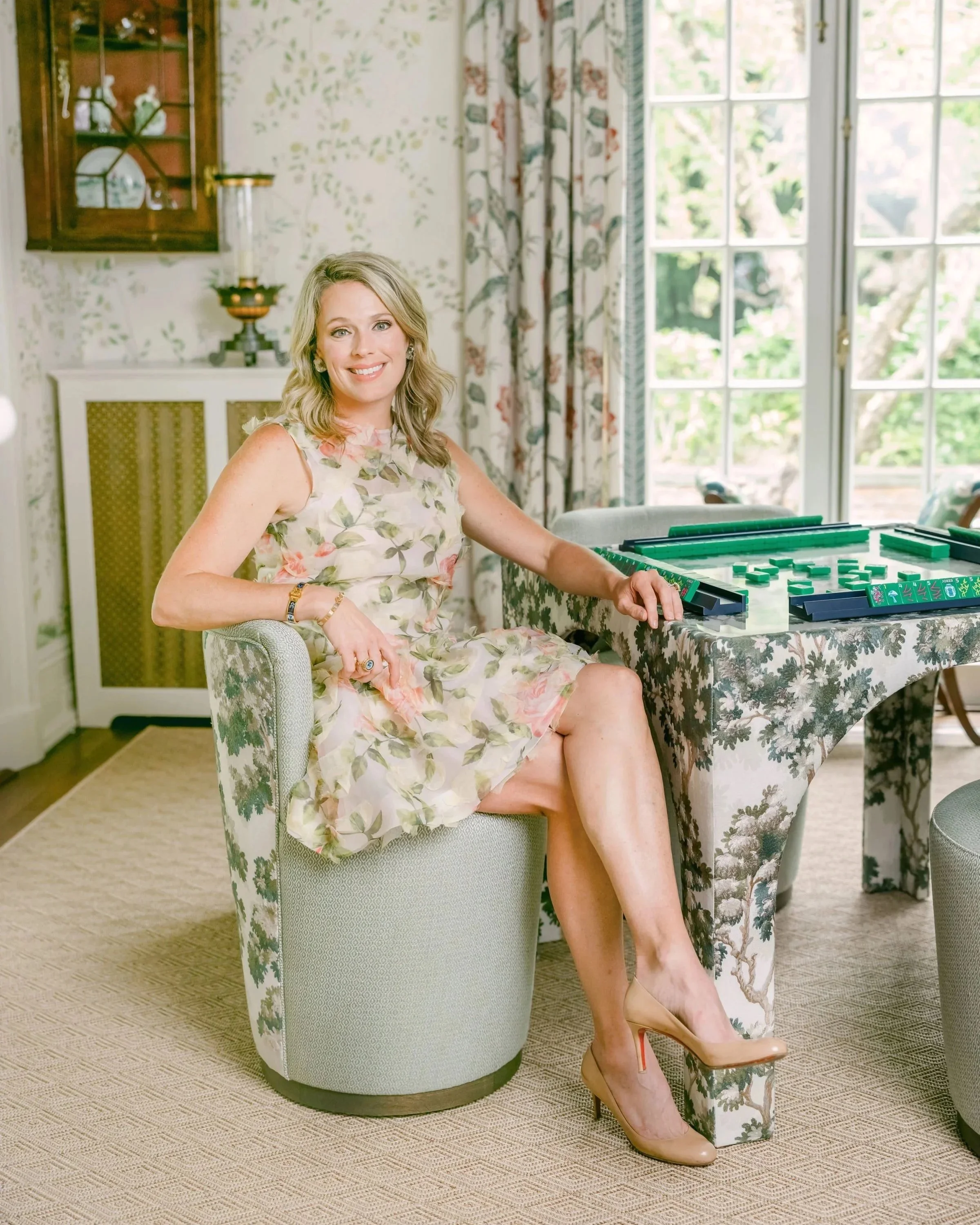 A woman in a floral dress, beige high heels, and jewelry sitting on a green armchair in a brightly lit room. She is smiling at the camera, with one leg crossed over the other. The room features a table with a pattern tablecloth, a large window with curtains, and a sideboard with a lamp and decorative items.