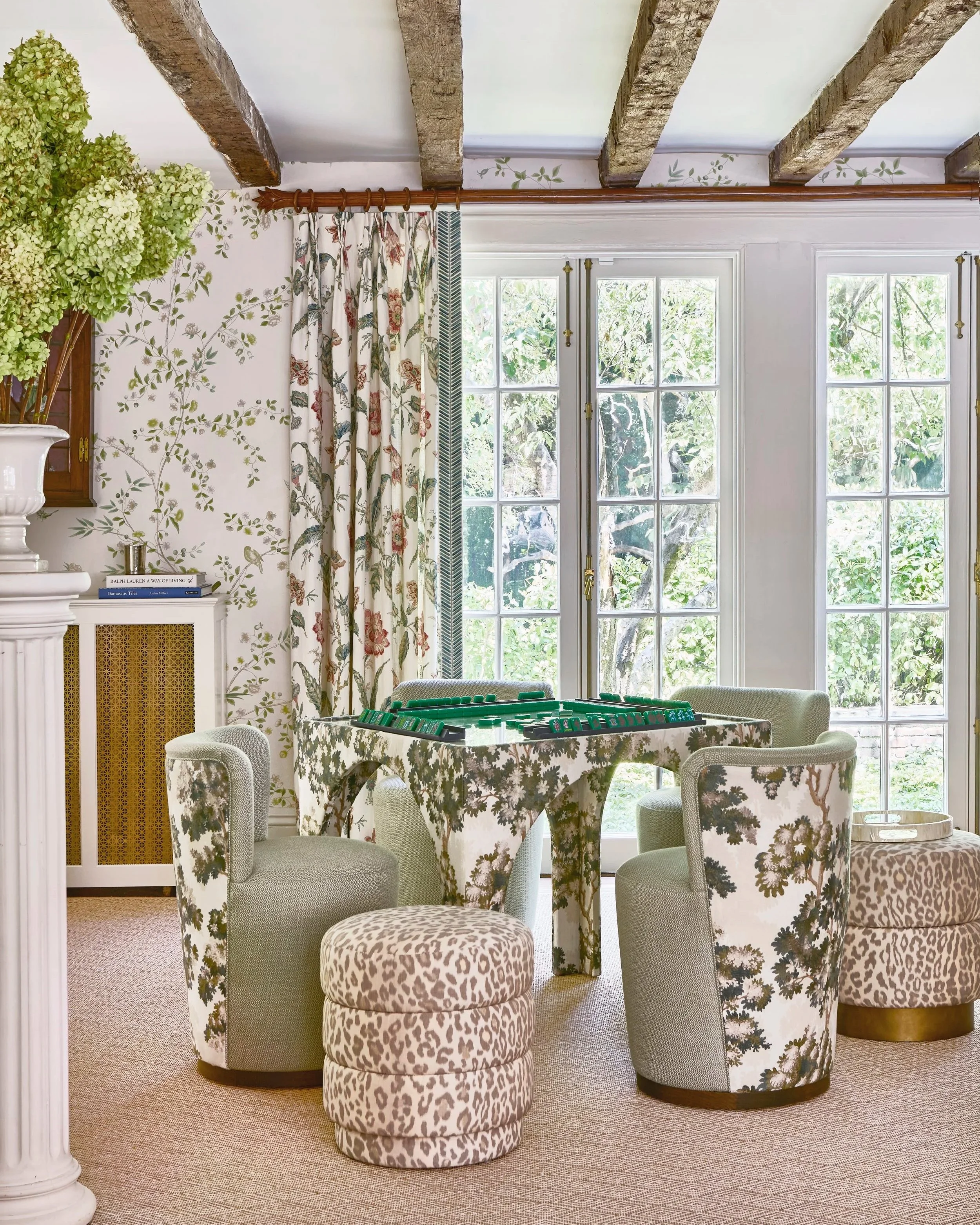 A round table with a floral tablecloth surrounded by six upholstered chairs, a small group of layered stools with animal print, large windows with white framing and brass hardware behind, and floral patterned curtains in a bright room with natural light.