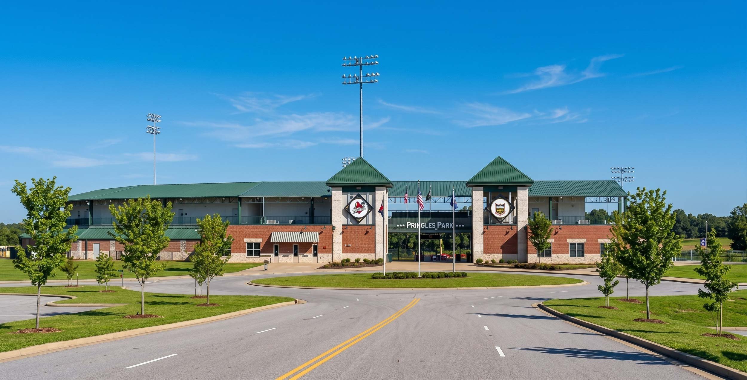 Front view of Pringles Park, a stadium with a brick and stone facade, green roof, and flags flying in front. The parking lot and trees are in the foreground, and the sky is clear with a few clouds.