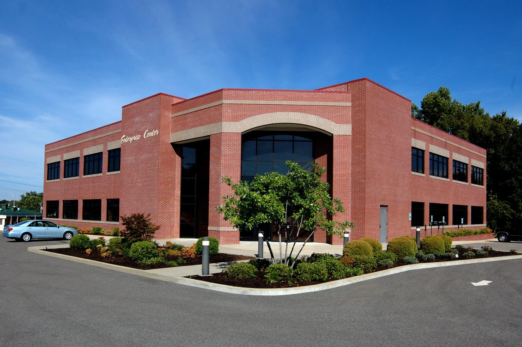 A two-story red brick commercial building with an arched entrance and large windows, surrounded by landscaped bushes and trees, with a parking lot in front and blue sky overhead.