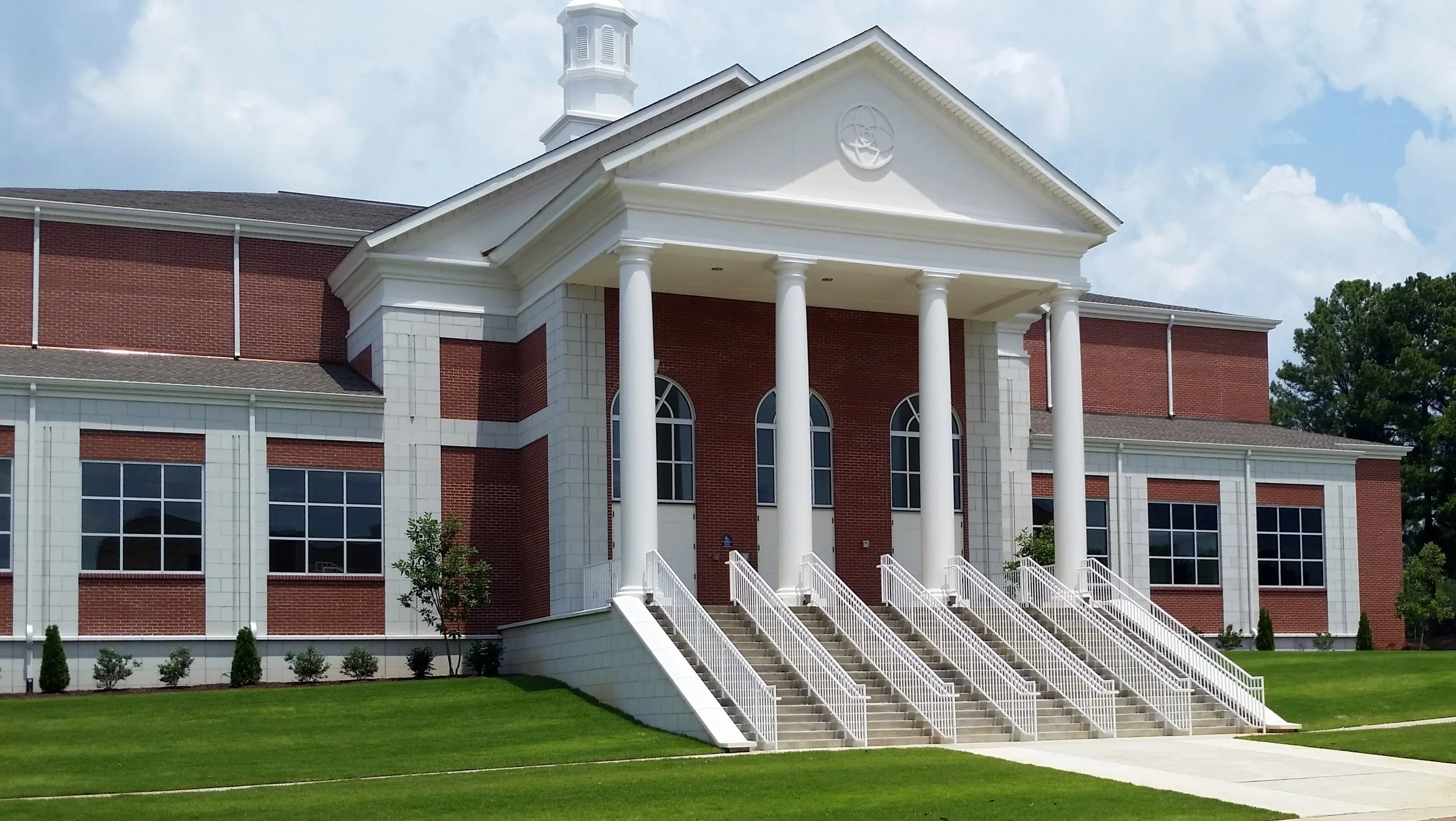 Front view of a church building with red brick walls, white columns, and a staircase leading up to the entrance, under a partly cloudy sky.