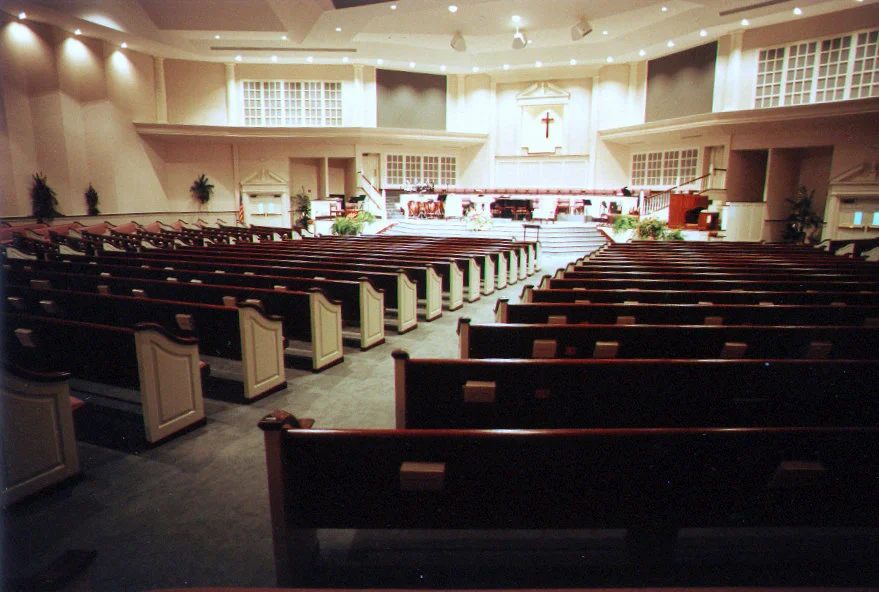Interior of a church sanctuary with wooden pews leading up to the altar, which has a cross on the wall behind it, and seating areas with musical instruments and microphones.