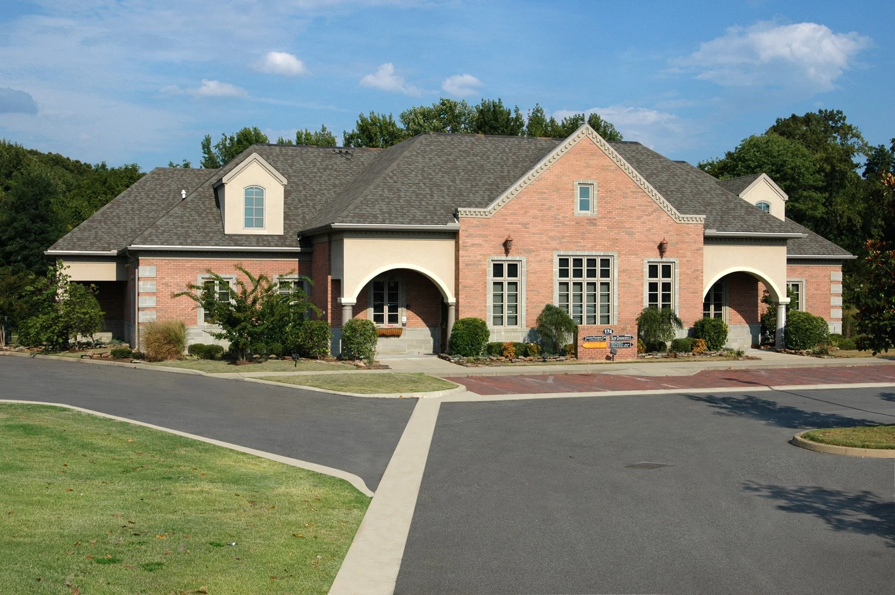 A large, two-story house with a brick and stucco exterior, multiple windows, and a curved driveway in front, surrounded by small landscaped bushes and trees, with a clear blue sky in the background.