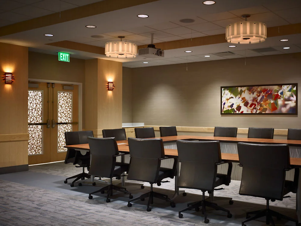 Empty conference room with a large rectangular table, black office chairs, a framed artwork of colorful leaves on the beige wall, wooden double doors with decorative glass, green exit sign, ceiling lights, and wall-mounted lamps.