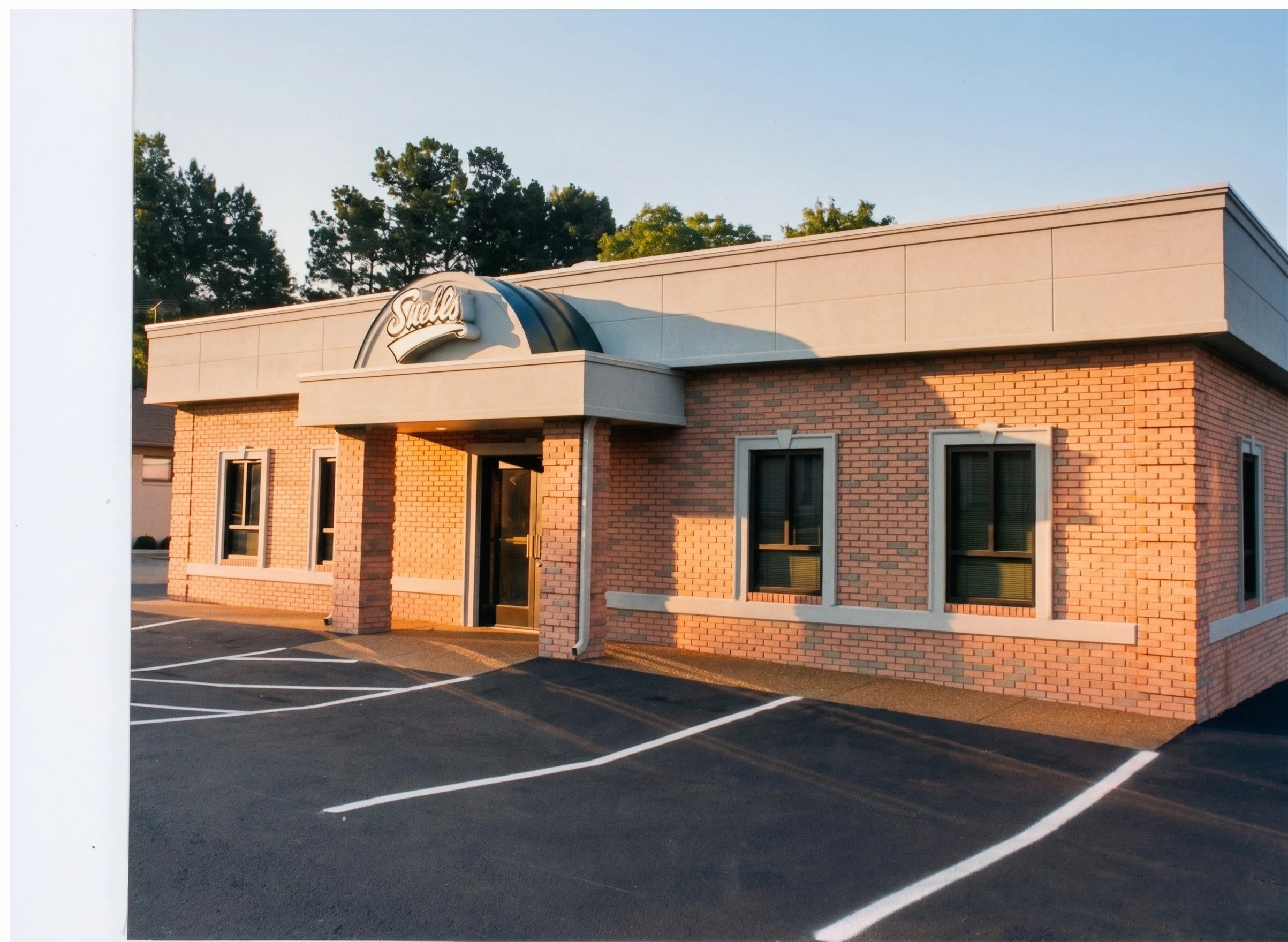 A brick commercial building with a sign that reads 'Stella' on the roof, surrounded by a parking lot with empty parking spaces and marked lines, with trees in the background.