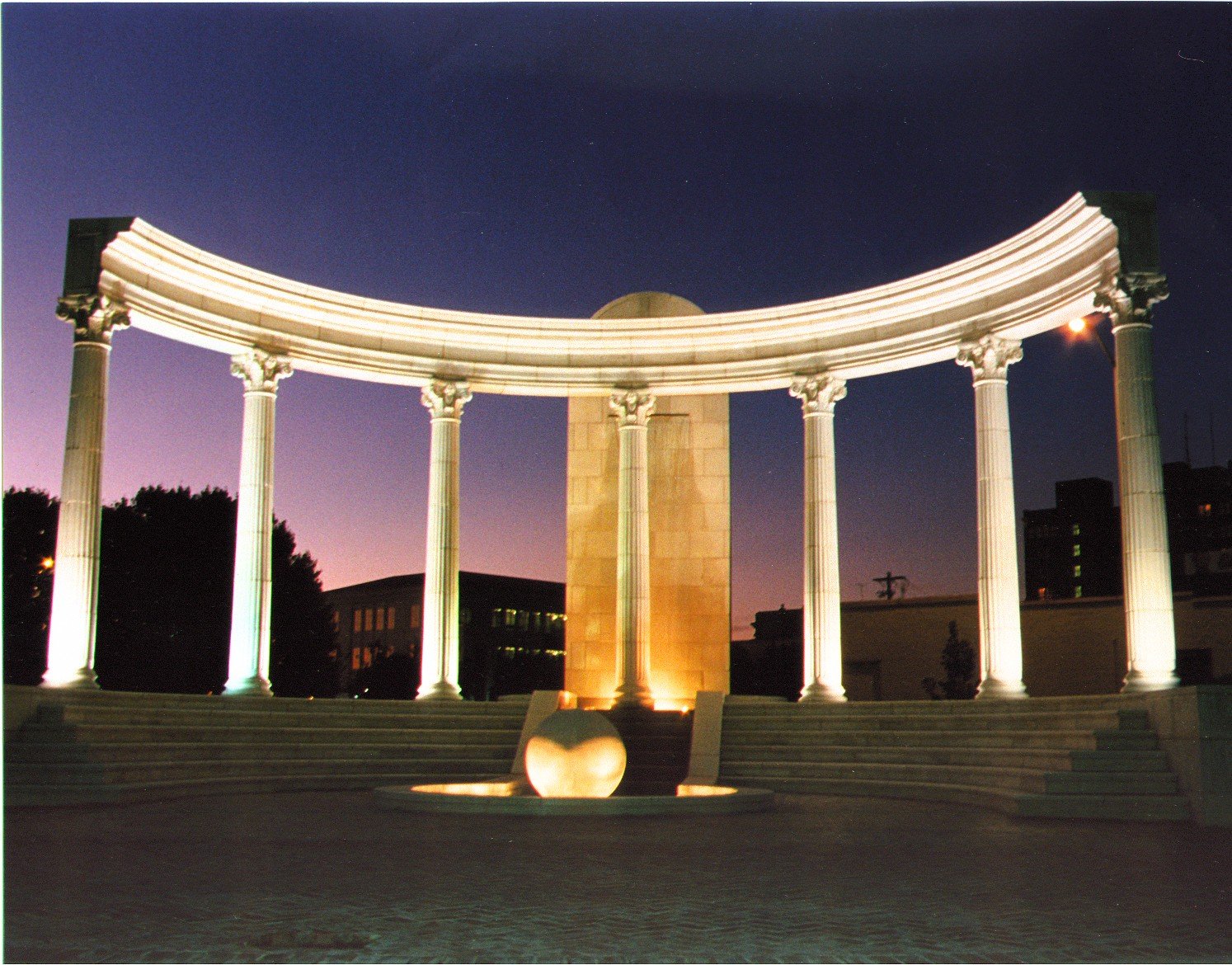 A monument with a round collumns and a curved roof, illuminated at night with pink and purple sky in the background