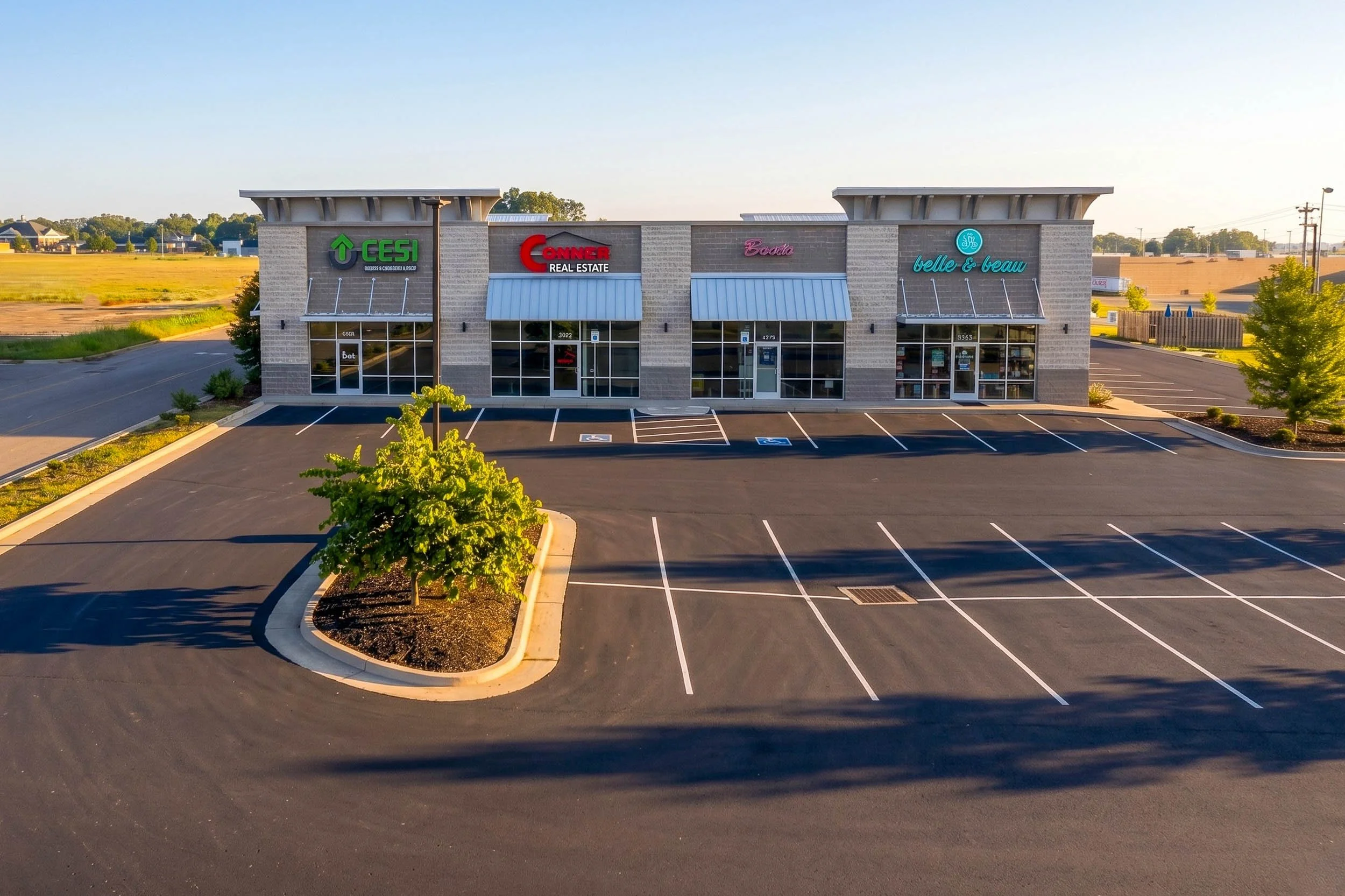 Empty parking lot in front of a small shopping center with four stores: CESI, Conner Real Estate, Booda, and Belle & Beaux, surrounded by trees and open land during daylight.